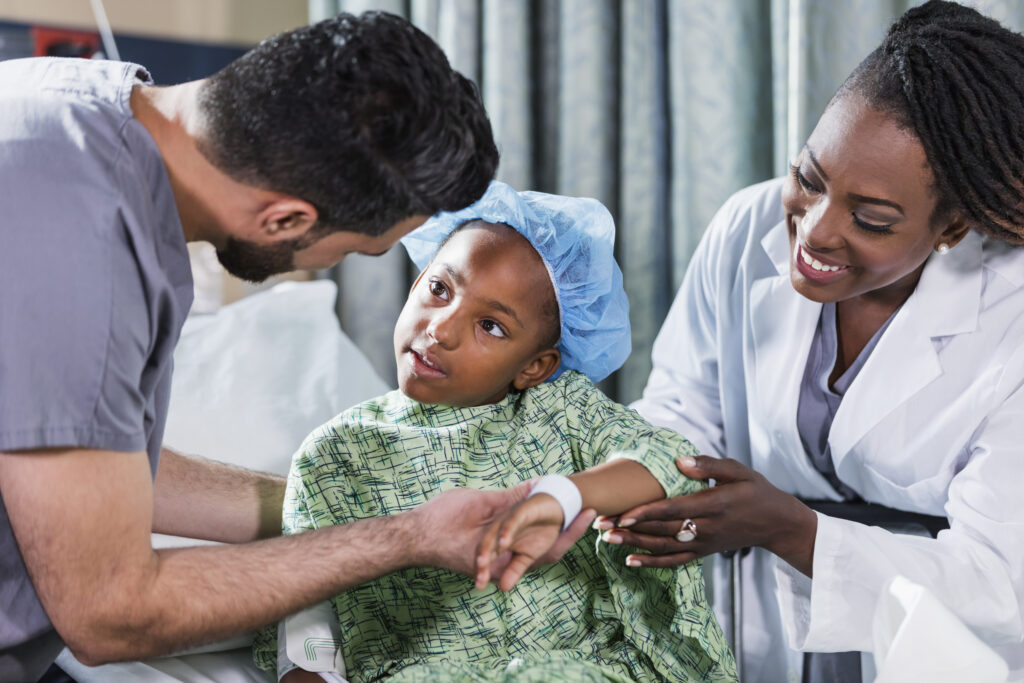 Una bambina afro-americana di 6 anni è seduta su una barella in ospedale, assistita da un medico e un’infermiera. Indossa un camice ospedaliero e una cuffia chirurgica. Un infermiere in divisa chirurgica in sala operatoria, un giovane uomo mediorientale sui 20 anni, sta in piedi da un lato e una dottoressa, una donna afro-americana sui 40 anni, sta in piedi dall’altro lato.